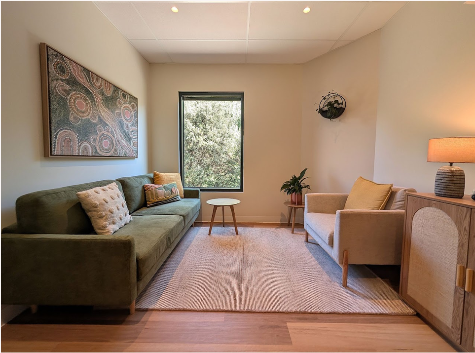 A consulting room at Hawthorn Hub with a green sofa, upholstered armchair, wall artwork and a leafy window outlook